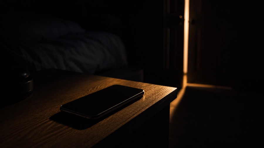 A phone lying dark and silent on a bedside table at night, lit only by a sliver of light from the doorway.