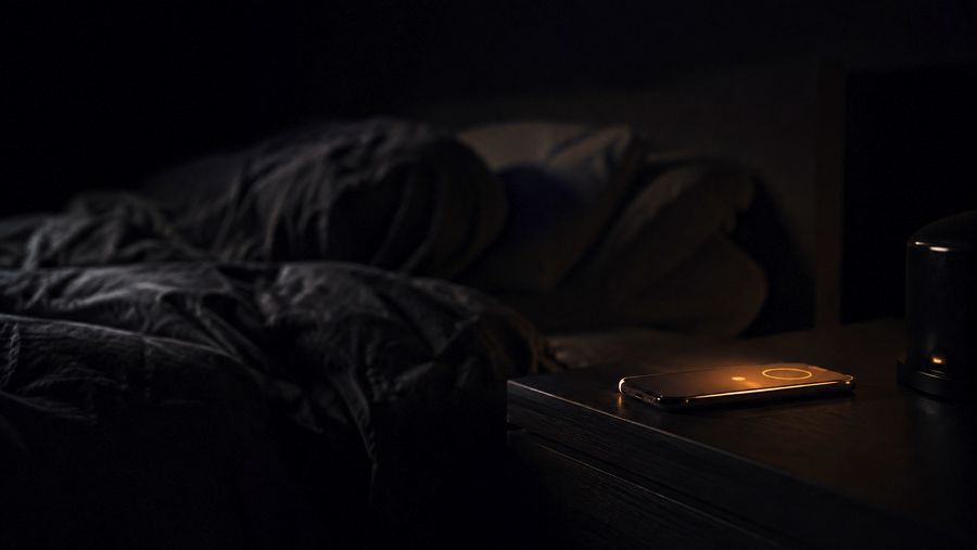 A sleeping figure under a duvet in near-darkness while a phone glows on the bedside table, unheard.